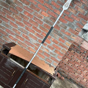 May include: A silver spear with a decorative blade featuring an ornate design. The spear has a black grip and a rounded end. The background is a weathered brick wall and a dark brown cabinet.