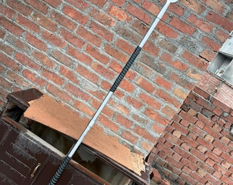 May include: A silver spear with a decorative blade featuring an ornate design. The spear has a black grip and a rounded end. The background is a weathered brick wall and a dark brown cabinet.