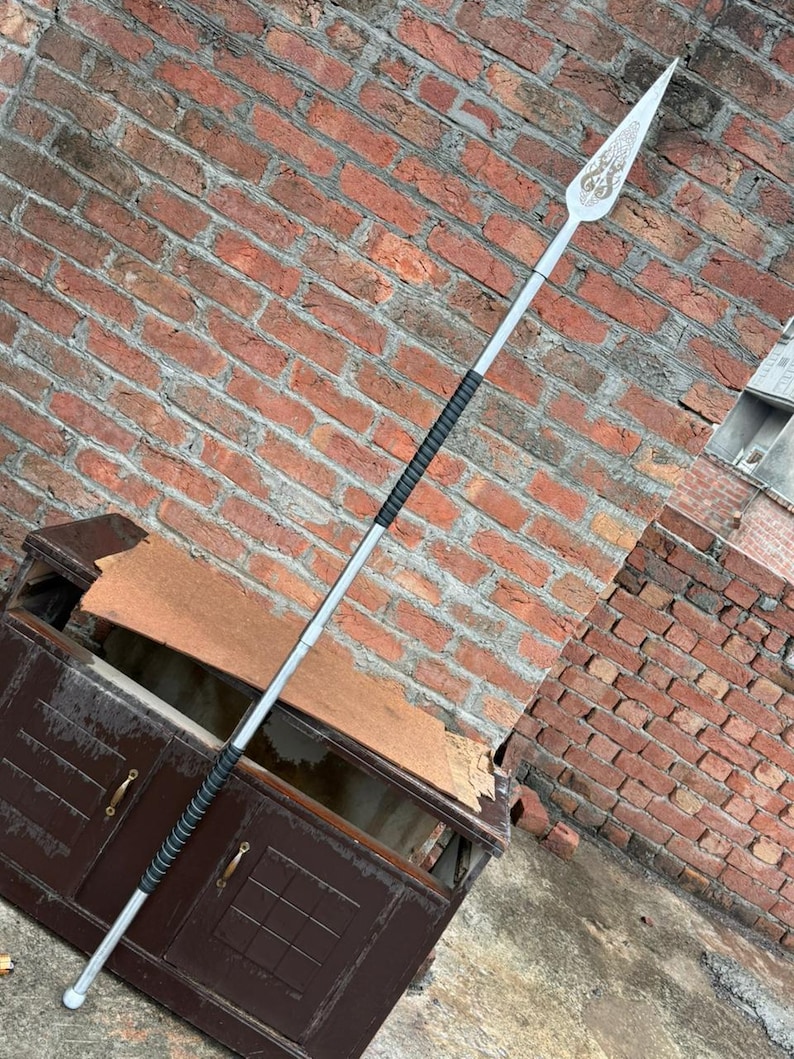 May include: A silver spear with a decorative blade featuring an ornate design. The spear has a black grip and a rounded end. The background is a weathered brick wall and a dark brown cabinet.