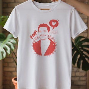 Puede incluir: Camiseta blanca con una ilustración roja de un hombre sonriente, el texto "Papi" y "Pedro Pascal", un corazón y una corona de laurel. La camiseta está colgada en una percha de madera.
