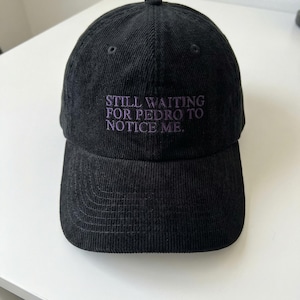 May include: Black corduroy baseball cap with the embroidered text "STILL WAITING FOR PEDRO TO NOTICE ME" in purple. The cap has a curved brim and a structured crown. A casual accessory.