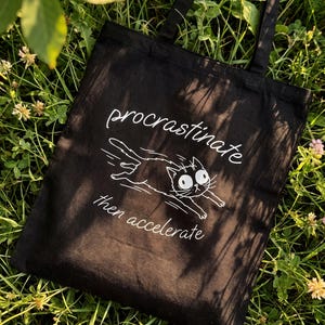May include: A black tote bag featuring a white graphic of a wide-eyed cat running with motion lines. The text "procrastinate" is printed above the cat, and "then accelerate" is printed below the cat in a white, cursive-style font.