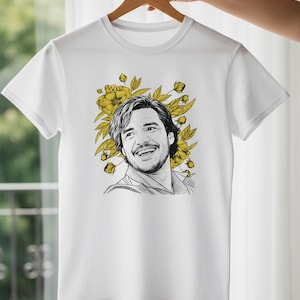 Peut inclure: T-shirt blanc avec un portrait en noir et blanc d'une personne souriante, entouré de fleurs et de feuilles jaunes. Le motif est centré, créant un contraste saisissant sur le tissu.