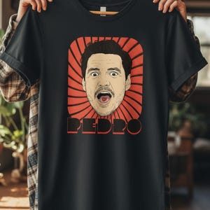 May include: Black t-shirt featuring a stylized illustration of a man's face with an open mouth and the name "PEDRO" in red block letters. The face is surrounded by a red burst design.