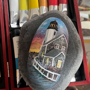 May include: A hand-painted rock depicting a lighthouse with a white exterior and a gray roof. The lighthouse is set against a colorful sunset sky with a blue sky above. The rock is gray and has a smooth surface.