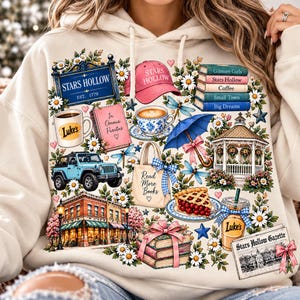 May include: A cream-coloured hoodie featuring a colourful graphic print of Stars Hollow elements including a blue sign, pink cap, stacked books, Luke's mug, a blue Jeep, a gazebo, a slice of cherry pie, a tote bag, and a Stars Hollow Gazette newspaper.