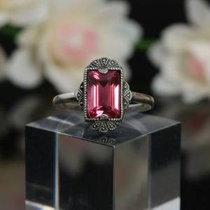 Può includere: Un anello d'argento con una pietra preziosa rosa rettangolare. La pietra preziosa è incastonata in una cornice decorativa con piccoli accenti scuri. L'anello è esposto su un blocco quadrato trasparente.