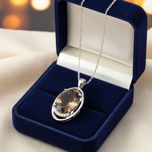 Può includere: Una collana d'argento con un grande pendente ovale in pietra preziosa marrone, esposta in un cofanetto di velluto blu scuro. Il pendente è incorniciato in argento e presenta piccole pietre trasparenti. Lo sfondo è sfocato con una calda luce dorata.