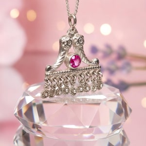 Op de afbeelding: Een zilveren hanger aan een ketting met een driehoekig frame van getextureerd metaal, een enkele ronde gefacetteerde roze edelsteen in het midden en een rij kleine bungelende cirkelvormige filigraanbedels aan de onderkant van een dunne zilveren ketting.