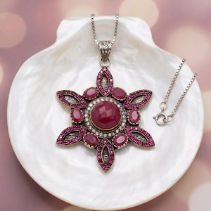 Pode incluir: Um colar com pendente em tom prateado com uma grande pedra central redonda, facetada e de cor rosa escuro, rodeada por pedras cor-de-rosa ovais mais pequenas e minúsculos cristais transparentes, com formato de estrela e metal texturizado.