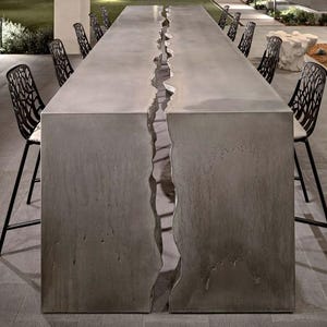 May include: A long, rectangular concrete table with a large crack running down the center. The table is surrounded by black metal chairs.