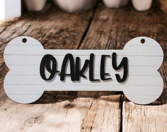 Könnte beinhalten: Weißer Holz-Hunde-Knochen mit dem Namen "Oakley" in schwarzen Buchstaben.