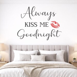 May include: A bedroom scene featuring a bed with white bedding and gray pillows. The wall above the bed has the text "Always KISS ME Goodnight" in black script, with a red lip print. Two bedside tables with lamps flank the bed.
