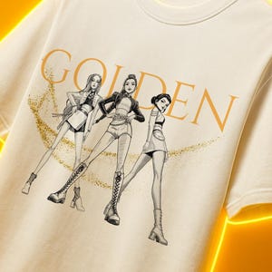 May include: Cream-colored t-shirt featuring a stylized illustration of three figures with the word "GOLDEN" in gold lettering. The figures are depicted in a fashion-forward style, with detailed clothing and accessories. A golden glitter effect surrounds the figures.
