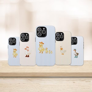 May include: Five phone cases featuring vertical stripes in blue, pink, and yellow. Each case displays a different illustration of ducks or ducklings wearing accessories like hats, boots, or bows, arranged in a row on a light-colored surface.