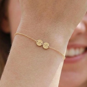 Op de afbeelding: Een fijne gouden armband met twee kleine, ronde bedels, elk gegraveerd met een enkele initiaal. De armband wordt om een pols gedragen, met een vage achtergrond.
