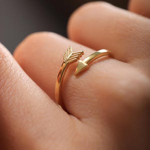 May include: A gold-coloured arrow ring. The ring has an open design with an arrow pointing towards the finger. The arrow's fletching and arrowhead are clearly defined. The ring is worn on a finger.