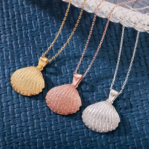 Può includere: Tre collane con ciondoli a forma di conchiglia in oro, oro rosa e argento. Ogni collana presenta un ciondolo a conchiglia strutturato e dettagliato, sospeso a una delicata catena. I ciondoli sono larghi circa 3,8 cm, mostrando un tema costiero.