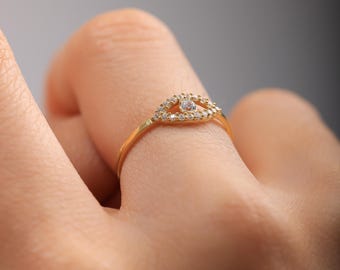 May include: A gold-colored ring featuring an eye-shaped design. The eye is outlined with small, clear gemstones, with a larger gemstone at the center. The ring is on a finger, with a blurred background.