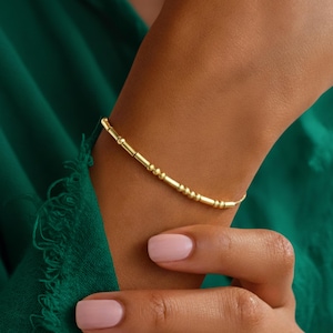 Könnte beinhalten: Ein goldfarbenes Armband mit filigranem Design. Das Armband besteht aus einer Reihe kleiner, runder Perlen und zylindrischen Segmenten, die aufgefädelt sind. Das Armband wird an einem Handgelenk getragen, im Hintergrund ist ein grünes Kleidungsstück zu sehen.