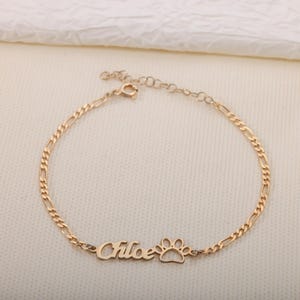 Può includere: Bracciale color oro con design a catena Figaro. Il bracciale presenta il nome "Chloe" in corsivo, seguito da un ciondolo a forma di zampa. Il bracciale è esposto su una superficie color crema.