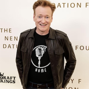 May include: A man wearing a black t-shirt with a microphone graphic and the word "HOME" inside a white circle. He is also wearing a brown leather jacket and blue jeans. The background is a white wall with text.