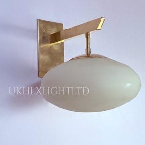 May include: A wall-mounted light fixture with a brass finish. The fixture features a rectangular backplate, a horizontal arm, and a white, oval-shaped glass shade. The text "UKHLXLIGHTLTD" is printed on the shade.