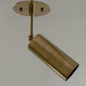 May include: A brass-colored, cylindrical spotlight fixture. The light is mounted to the ceiling with a round plate and a straight rod. The cylindrical light can be angled. The fixture is made of metal.