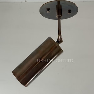 May include: A bronze-coloured, adjustable spotlight fixture. The cylindrical light is attached to a round ceiling plate via a rod and a swivel joint. The text "UKHLX LIGHTLTD" is printed on the light.
