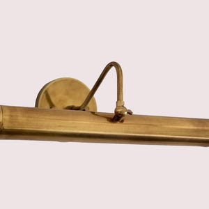 May include: A brass picture light with a cylindrical body and a curved arm. The light is mounted on a round base. The finish is a warm, golden colour, suggesting an antique or vintage style. The design is simple and functional.