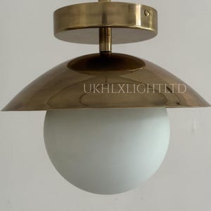 May include: A modern ceiling light fixture with a brass finish. The fixture features a round, white globe light and a brass shade with the text "UKHLXLIGHTLTD". The light is suspended from a brass ceiling mount.