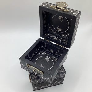 May include: Three stacked, open black wooden boxes with silver hardware. The boxes have a decorative white border and interior with a skull design. The top box is open, revealing the interior design. The boxes are on a white surface.