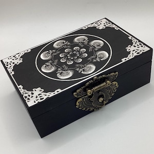 May include: Black wooden box with a decorative silver and skull design on the lid. The box features a metal clasp and ornate silver corner accents. The skull design is arranged in a circular pattern.