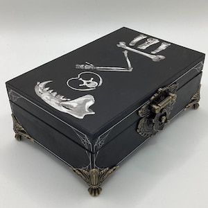 May include: A black wooden box with a hinged lid, featuring a decorative metal clasp and corner feet. The lid is adorned with white illustrations of bones and a heart shape. The box has a gothic or Halloween theme.