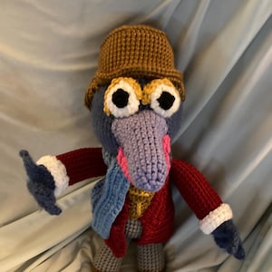 Peut inclure: Poupée au crochet faite à la main, corps bleu, chapeau marron, veste rouge et écharpe bleue. La poupée a de grands yeux bordés de blanc et un bec violet. Les bras et les jambes sont ornés de poignets blancs et de bottes marron.