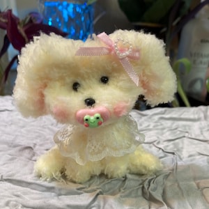 May include: A small, cream-colored plush toy dog with a pink bow and a green frog pacifier. The dog is wearing a white lace collar.