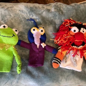 May include: Three crocheted hand puppets: a green frog, a purple bird with a blue head, and an orange and red monster with a white shirt.