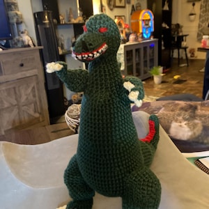 May include: Green crocheted dinosaur toy with red accents and white claws. The dinosaur has a menacing expression with red eyes and sharp teeth.