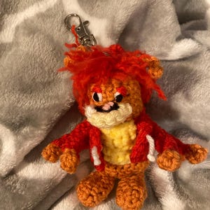May include: Crocheted keychain featuring a small, orange lion with a red mane and jacket. The lion has a yellow chest and white accents. The keychain has a silver clasp.