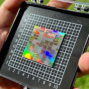 Può includere: Un chip per computer in silicio quadrato con una finitura iridescente arcobaleno, centrato su un vassoio a griglia in plastica nera contrassegnato con le lettere da A a J e i numeri da 1 a 10. Il chip presenta intricati motivi di circuiti.