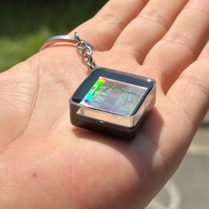 May include: A square keychain featuring a central computer processor chip encased in a clear, glossy resin block with a black base. The chip displays iridescent, rainbow-colored light reflections across its intricate metallic circuitry patterns.