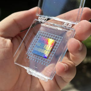 Pode incluir: Um pequeno chip de processador de computador quadrado e iridescente está dentro de uma caixa protetora de plástico transparente. A caixa apresenta uma grade impressa em branco com letras de A a J e números de 1 a 10 nas bordas.