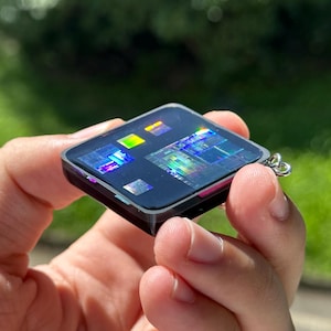 Puede incluir: Un llavero de forma cuadrada con una superficie oscura y brillante incrustada con múltiples chips de procesador de computadora iridiscentes y multicolores. Los chips muestran un patrón de rejilla holográfico reflectante con tonos vibrantes.