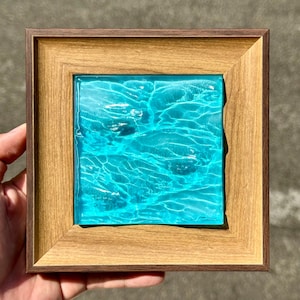 Può includere: Una cornice quadrata in legno con un bordo esterno scuro e un bordo interno più chiaro, che racchiude un inserto in vetro turchese traslucido e testurizzato che imita l'aspetto dell'acqua increspata con riflessi di luce e ombre.