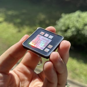 Può includere: Un portachiavi quadrato con cornice nera e un microchip incorporato al centro. Il chip mostra intricati motivi circuitali multicolori con una finitura iridescente riflettente che cattura la luce, creando un bagliore vibrante.