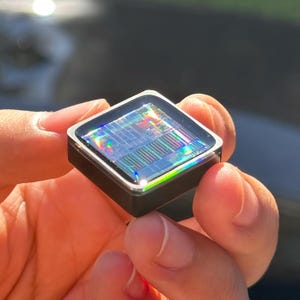 Könnte beinhalten: Eine kleine, quadratische elektronische Komponente mit einer dunklen, reflektierenden Oberfläche, die bei Lichteinfall komplexe, irisierende, regenbogenfarbene Schaltkreismuster zeigt. Das Objekt hat einen glatten, schwarzen Rahmen und eine flache, glänzende Oberseite.
