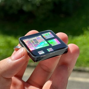 Può includere: Un portachiavi di forma quadrata con una superficie nera lucida incastonata con un microchip iridescente e colorato. Il circuito interno mostra una complessa griglia di motivi metallici che riflettono vivaci tonalità arcobaleno.