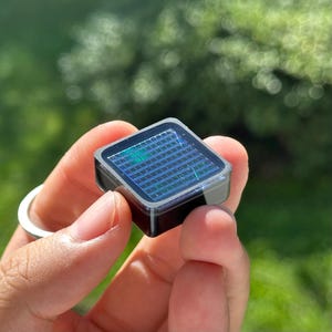 Op de afbeelding: Een kleine, vierkante, zwarte elektronische component met een rasterachtig zonnepaneeloppervlak met een reflecterende, iriserende blauwe en groene afwerking. Het apparaat heeft een gladde, donkere behuizing en een transparante toplaag.
