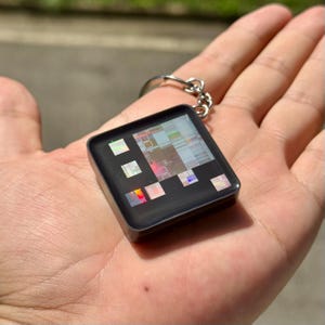 以下が含まれることがあります： 黒いフレームと光沢のある表面を持つ正方形のキーホルダー。表面にはホログラフィック効果を示す小さな虹色の多色長方形チップが埋め込まれており、銀色の金属製二重リングに取り付けられています。