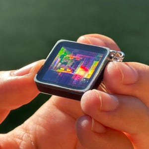 Puede incluir: Un colgante cuadrado para llavero con un marco oscuro y brillante y una incrustación central que muestra un patrón de placa de circuito de microchip vibrante y multicolor con tonos iridiscentes de rojo, verde, amarillo y azul, con una anilla de metal plateado.